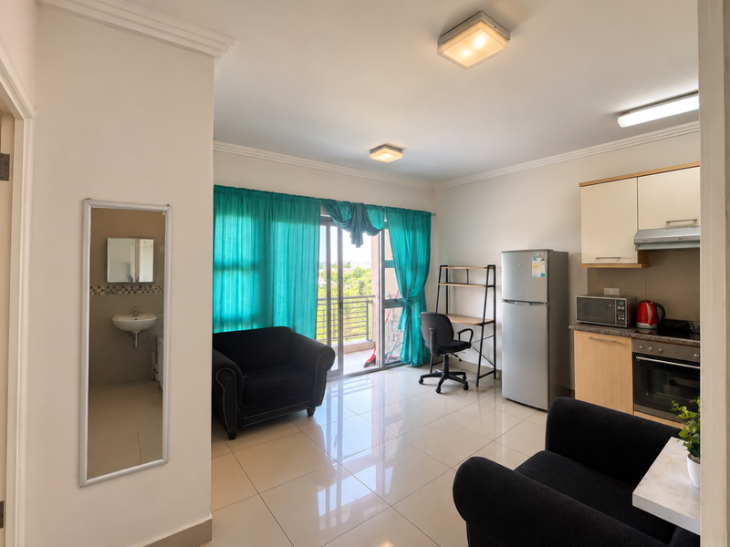 Open-plan living area featuring tiled floors, a compact kitchen with built-in appliances, and direct access to a balcony through sliding glass doors.