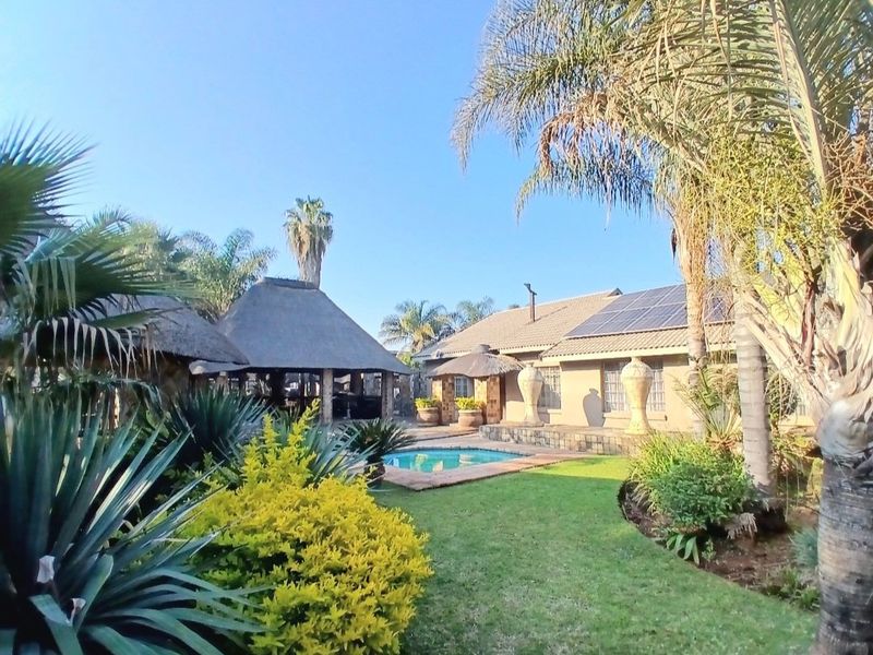 A spacious rear garden featuring a swimming pool, a traditional thatch roof lapa, and a main house equipped with solar panels.