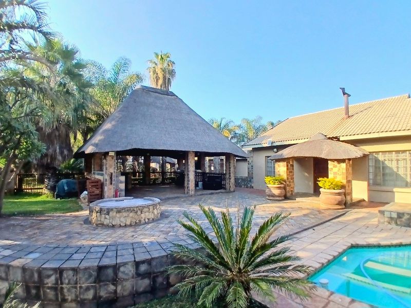 Spacious outdoor entertainment area featuring a large thatched lapa, paved patio, stone fire pit, and an adjacent swimming pool.