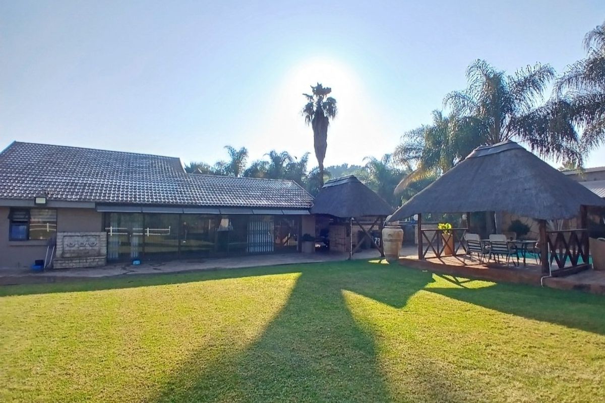Spacious rear garden featuring a well-maintained lawn, a large tiled-roof house with sliding doors, and a traditional thatched lapa for outdoor entertainment.