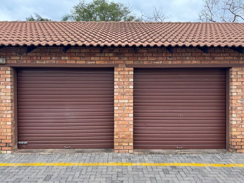 A double garage featuring brown roller doors, a face brick exterior, and a tiled roof, situated above a paved driveway.