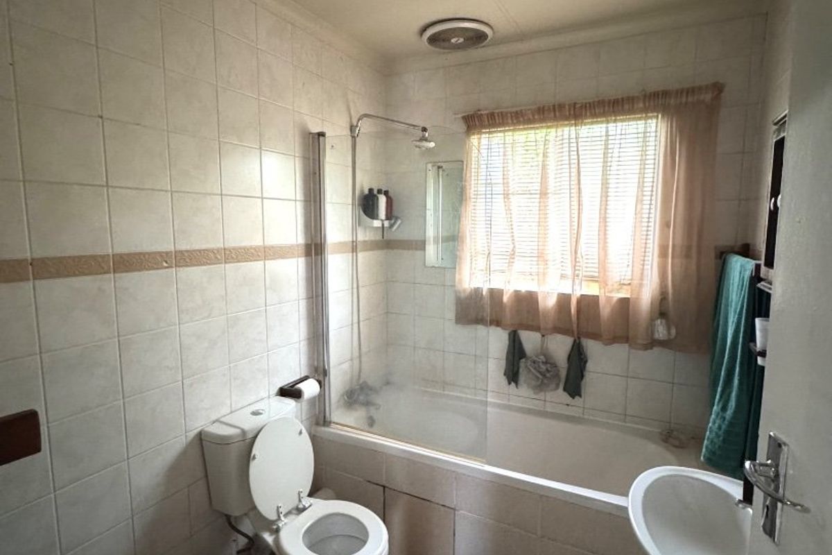 A dated bathroom featuring a bathtub with a shower screen, pedestal sink, and toilet, finished with neutral wall tiling and an extractor fan.