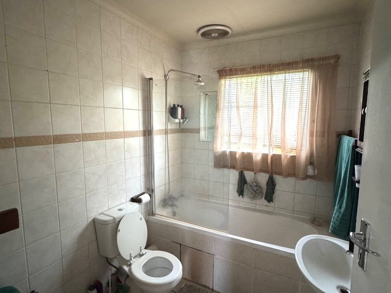 A dated bathroom featuring a bathtub with a shower screen, pedestal sink, and toilet, finished with neutral wall tiling and an extractor fan.