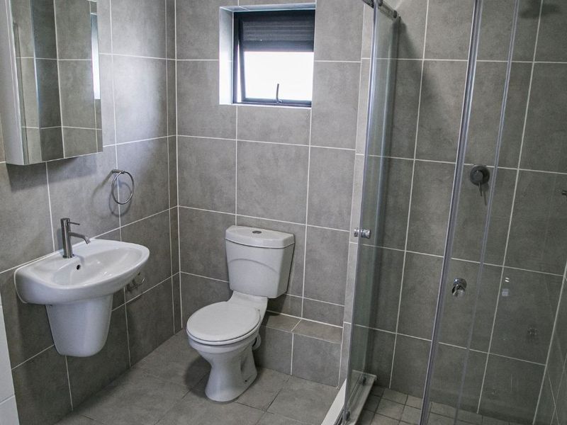 Modern bathroom featuring grey wall and floor tiles, a pedestal washbasin, a toilet, and a glass-enclosed shower.