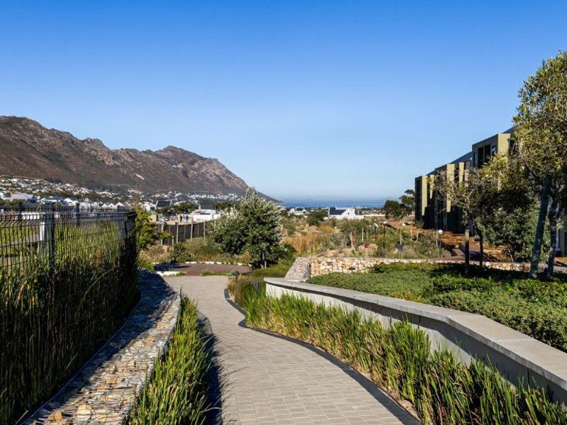 A scenic outdoor view featuring a paved walkway, landscaped gardens, and stone retaining walls, set against a backdrop of mountains and coastal landscape.