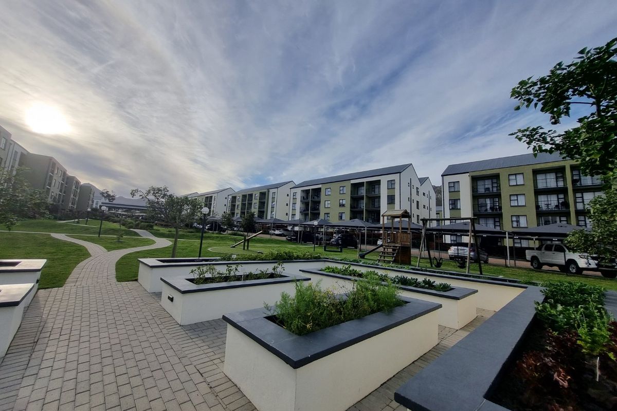 A well-maintained communal garden area featuring paved walkways, raised planter beds, and covered parking bays within an apartment complex.