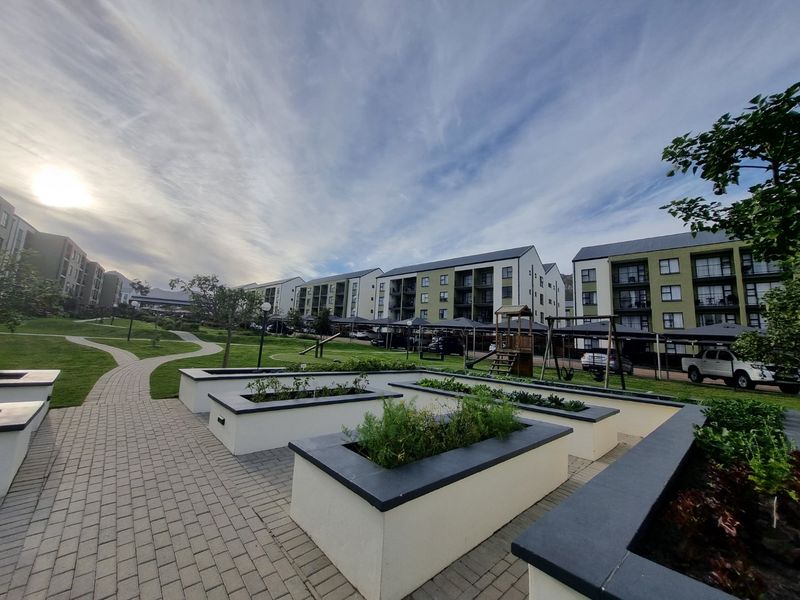 A well-maintained communal garden area featuring paved walkways, raised planter beds, and covered parking bays within an apartment complex.