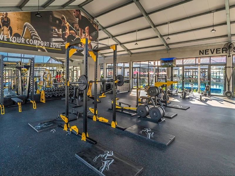 A well-equipped, modern gym facility featuring high ceilings with exposed steel trusses, large windows, and extensive mirrored walls for a spacious workout environment.