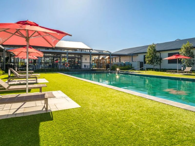 A well-maintained outdoor swimming pool area featuring a paved deck, artificial lawn, and surrounding communal building structures.