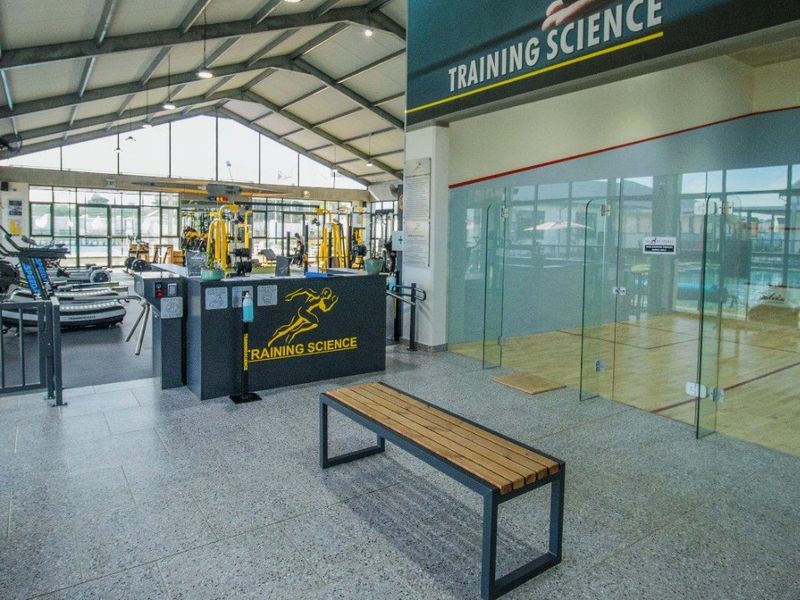 Modern sports facility featuring a glass-walled squash court, open-plan gym area with exposed steel roof trusses, and durable tiled flooring.