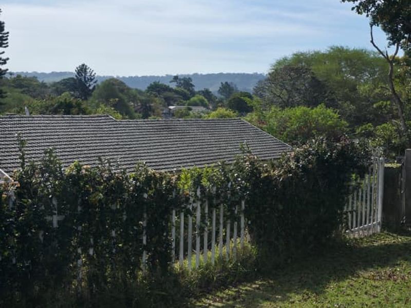 A scenic view overlooking a residential tiled roof and a white picket fence, set against a backdrop of lush greenery and distant hills.