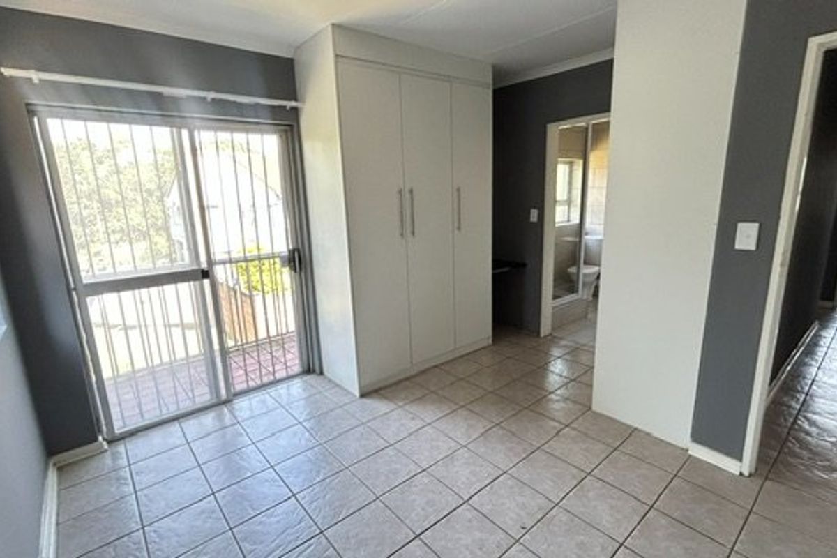 A bright, tiled bedroom featuring built-in cupboards, a sliding glass door with security bars, and direct access to an en-suite bathroom.