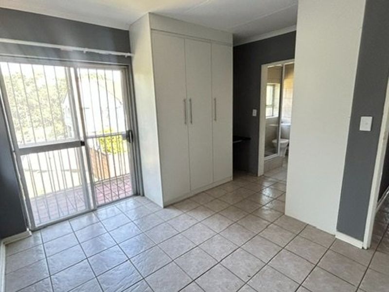 A bright, tiled bedroom featuring built-in cupboards, a sliding glass door with security bars, and direct access to an en-suite bathroom.