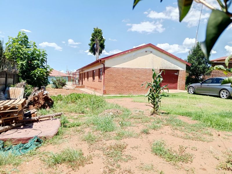 Rear view of a single-story brick house featuring a metal garage door and a large, unmaintained garden area.