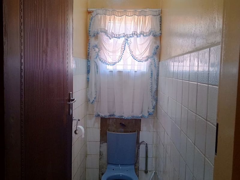 A small, dated bathroom featuring a blue toilet, partial wall tiling, and a window. The space requires renovation and cosmetic updates.