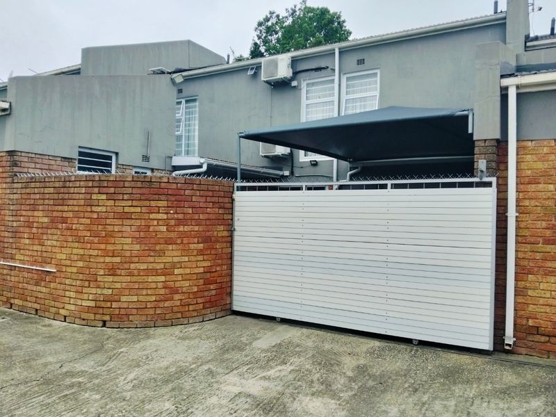 Exterior view of the townhouse featuring a secure sliding gate, brick boundary wall, and concrete driveway with an installed awning.