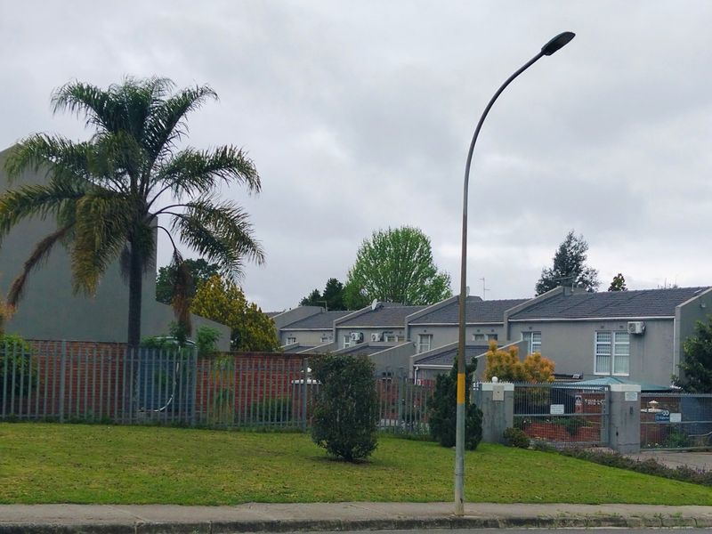 A street-level view of the townhouse complex, featuring grey exterior facades, tiled roofing, and secure perimeter fencing with gated access.