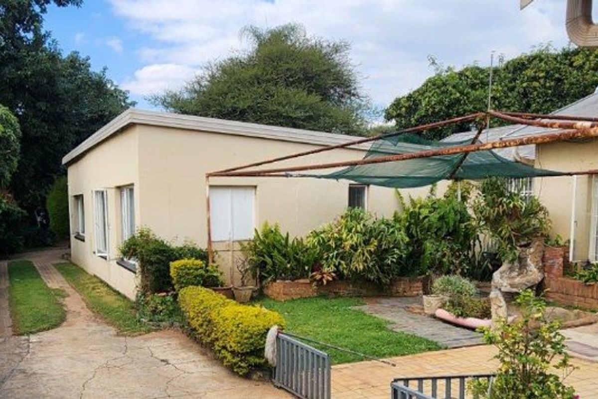 Rear exterior view showing a paved patio area, metal shade structure, concrete driveway, and established garden beds against the house facade.