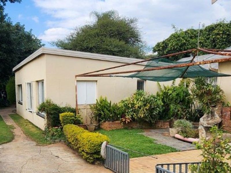 Rear exterior view showing a paved patio area, metal shade structure, concrete driveway, and established garden beds against the house facade.