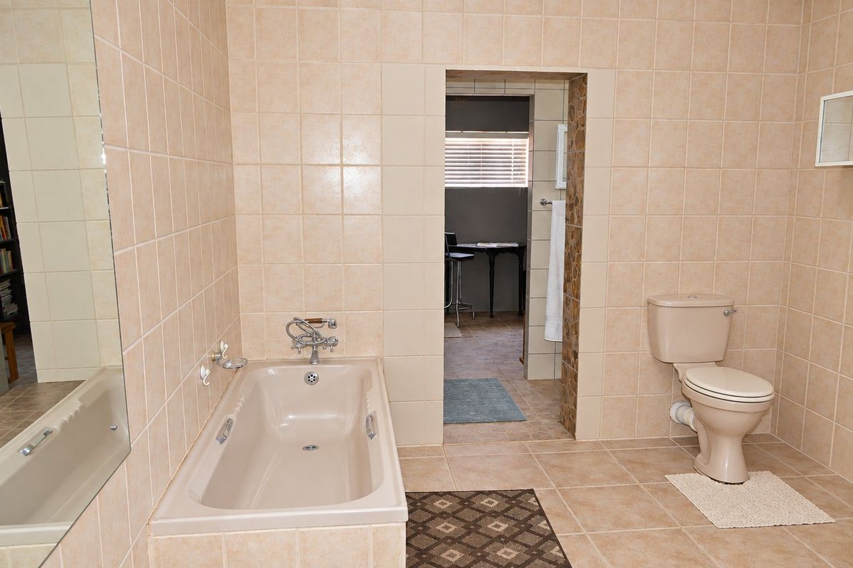A spacious bathroom featuring neutral wall and floor tiles, a built-in bathtub, and a standard toilet, with an adjoining room visible through the doorway.