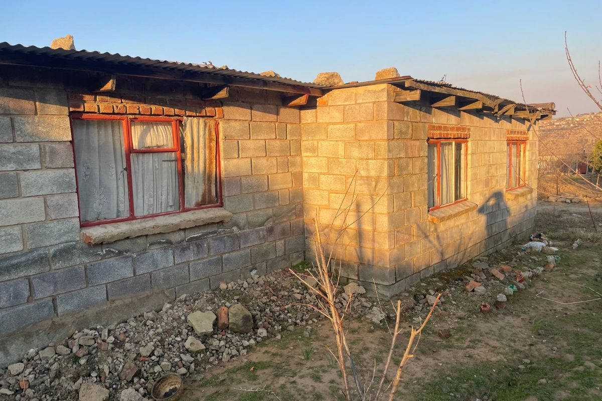 Exterior view of a single-story house featuring concrete block construction, steel-framed windows, and a corrugated metal roof in need of maintenance.