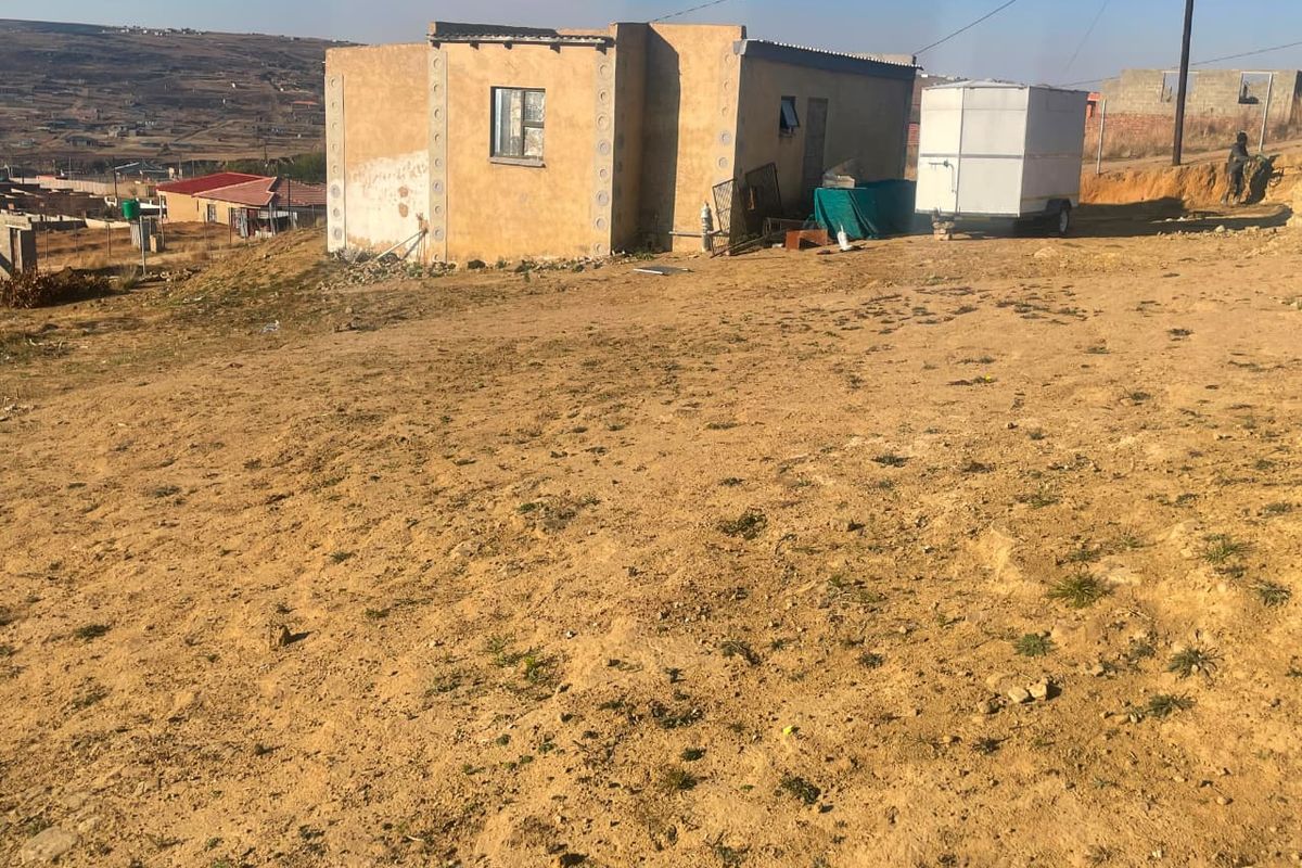 A detached single-story house featuring plastered masonry walls and a corrugated metal roof, situated on a large, undeveloped plot of land.