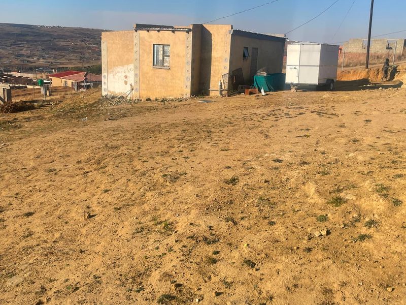 A detached single-story house featuring plastered masonry walls and a corrugated metal roof, situated on a large, undeveloped plot of land.
