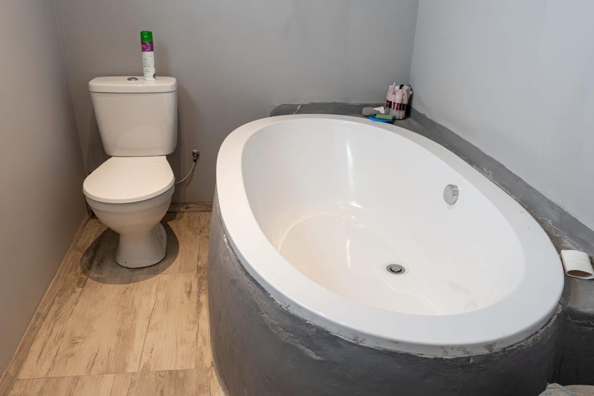 A bathroom featuring a built-in oval bathtub with a cement surround and a standard toilet, finished with wood-look floor tiles.