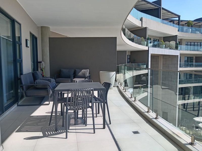 Spacious covered balcony featuring modern tiled flooring, a sleek glass balustrade, and direct access to the apartment through large sliding glass doors.