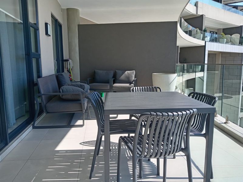 A spacious covered balcony featuring modern tiled flooring, a sleek glass balustrade, and direct access to the apartment interior through large sliding doors.
