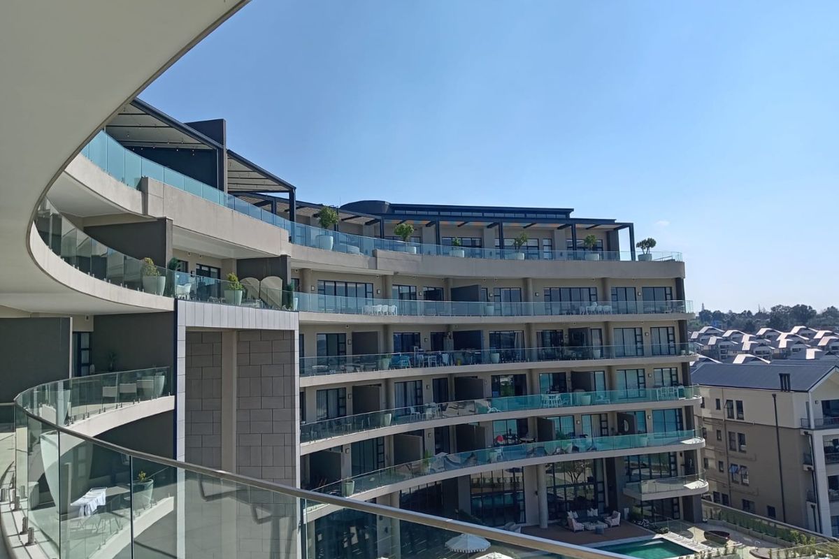 A view of a modern apartment complex featuring curved balconies with glass balustrades and a swimming pool area below.