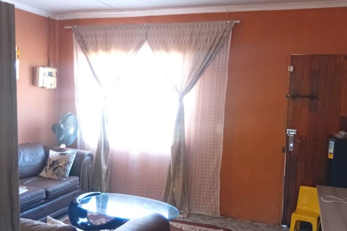 A living room featuring tiled flooring, a timber door, and an exposed electrical distribution board, with visible ceiling cracks and dated wall finishes.