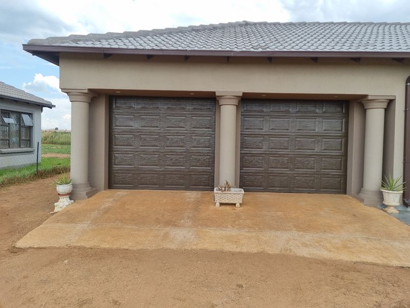 A double garage featuring dark panelled doors, decorative concrete columns, and a tiled roof, set against an unpaved driveway area.