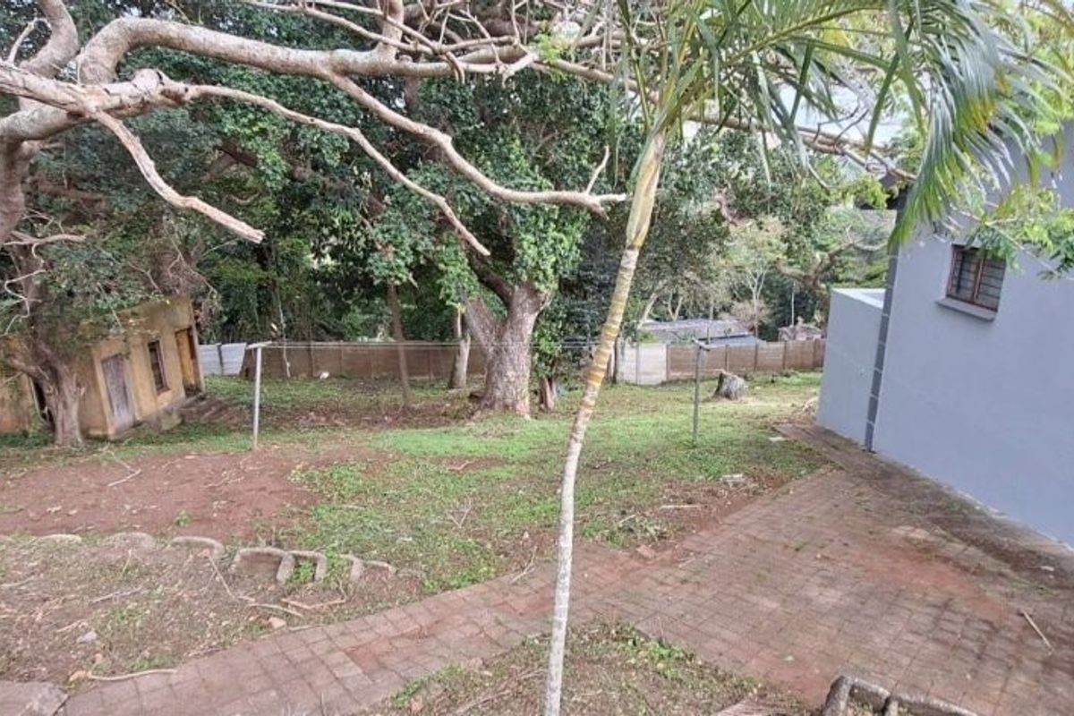 A view of the rear garden area featuring paved pathways, mature trees, and the side elevation of the house.