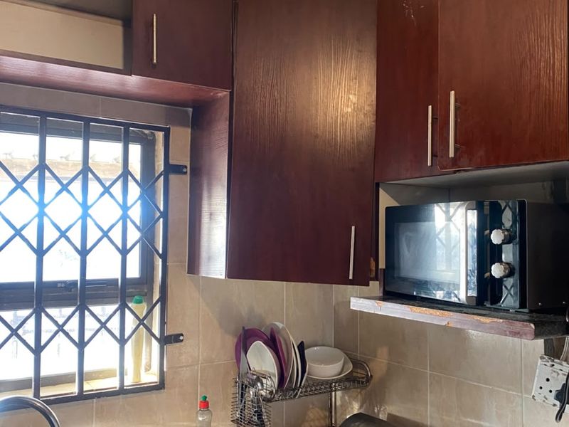 A compact kitchen featuring dark wood cabinetry, tiled backsplash, and a stainless steel sink, currently showing signs of wear and dated finishes.