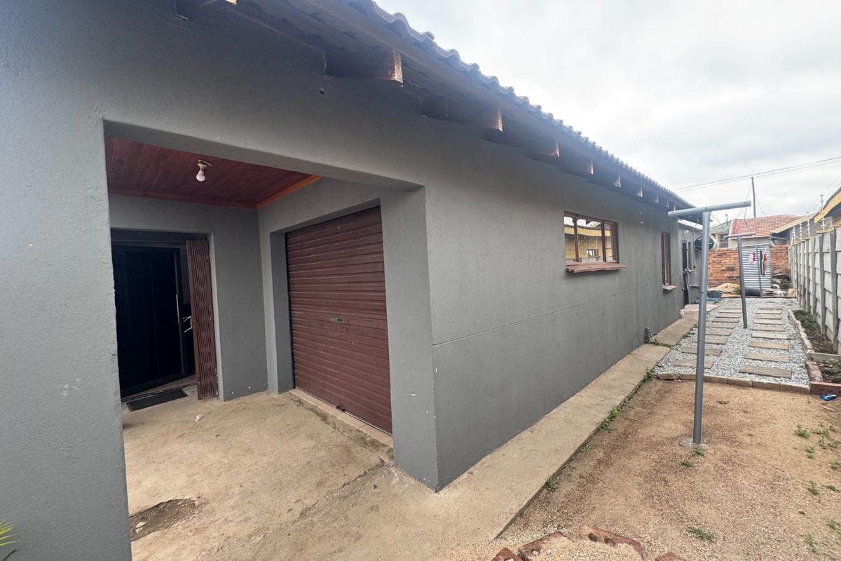 Rear exterior view showing a garage with a roller shutter door, concrete patio area, and a paved walkway leading along the side of the house.