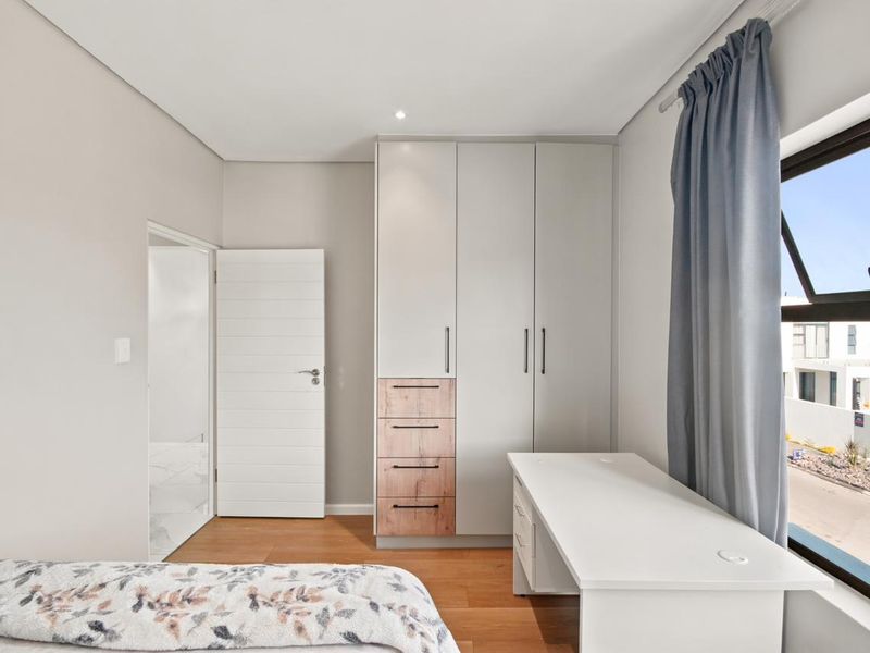 A modern bedroom featuring built-in cupboards with wood-accented drawers, laminate flooring, and a large window providing natural light.