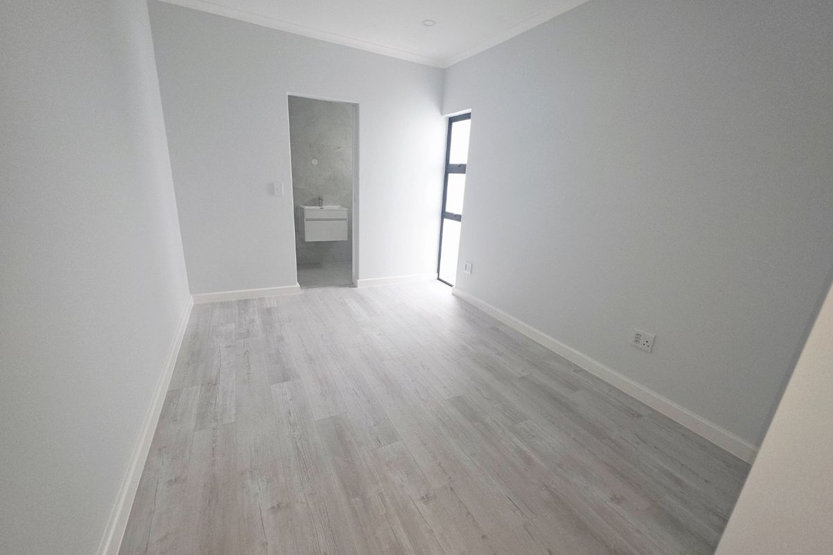 A bright, empty bedroom featuring modern light-toned laminate flooring, clean white walls, and an adjoining en-suite bathroom.
