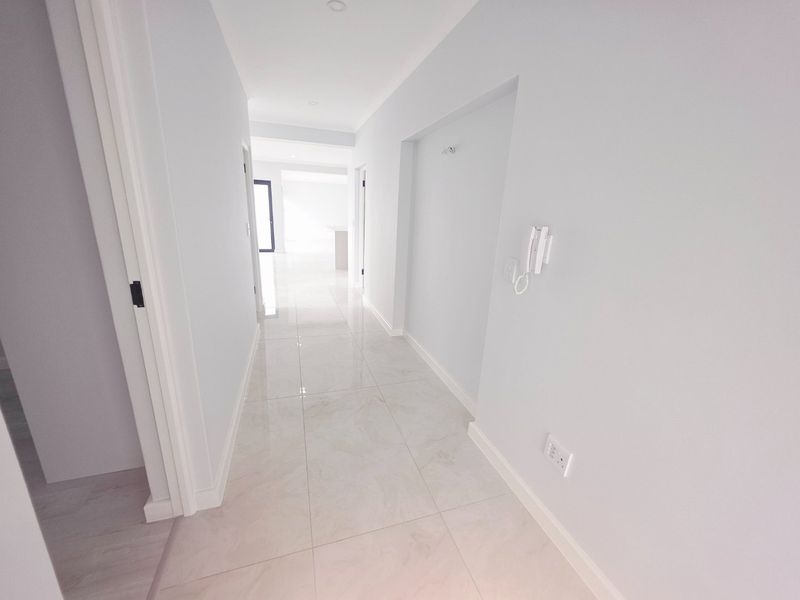 A bright, modern hallway featuring neutral tiled flooring, recessed lighting, and a wall-mounted intercom system, leading into an open-plan living area.