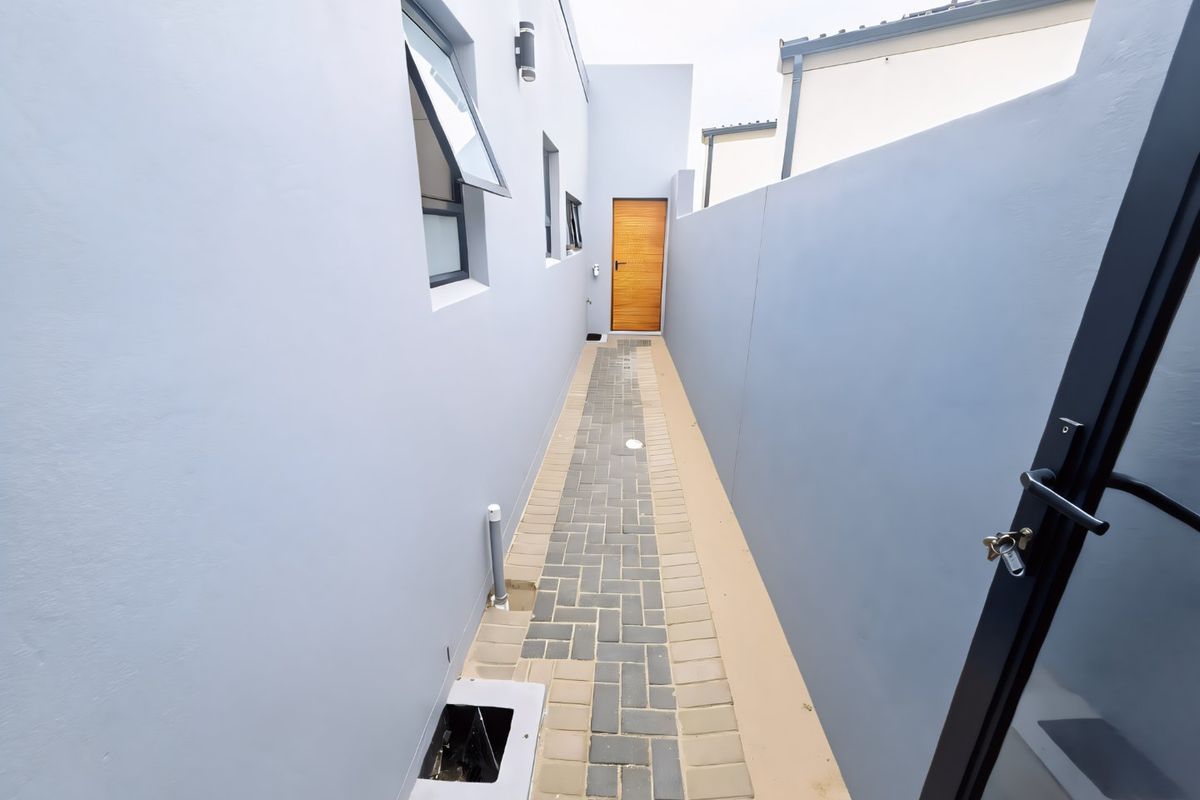 A clean, paved side passage or rear walkway featuring modern grey boundary walls, a timber door, and functional exterior lighting.