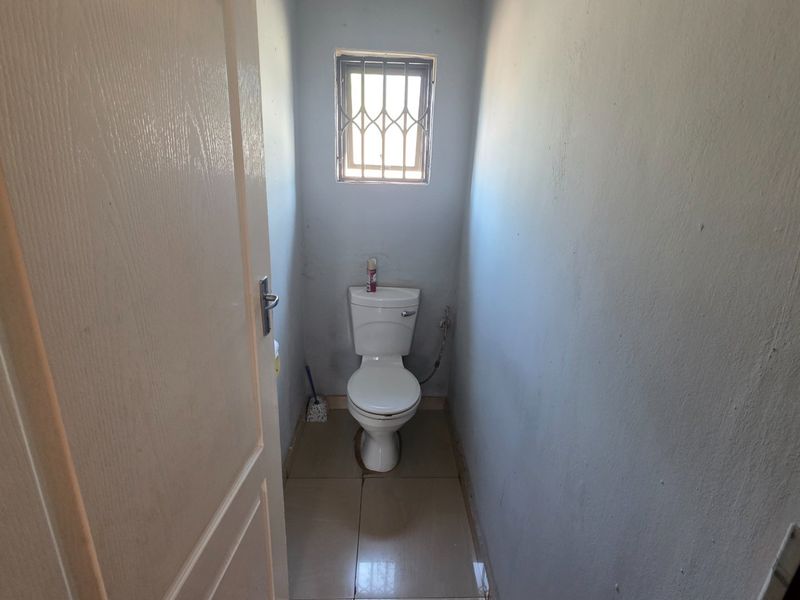 A small, functional guest toilet featuring tiled flooring and a security-barred window, currently in need of cosmetic updates.