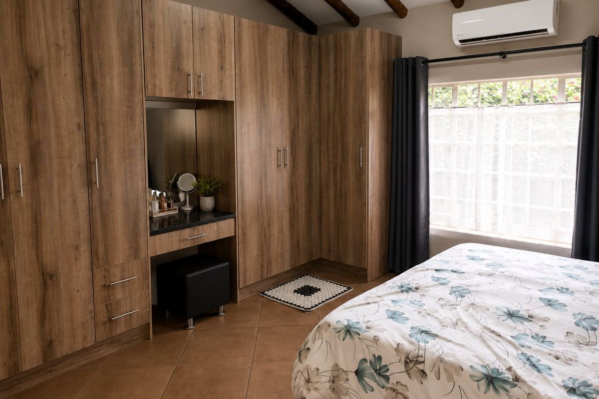 A well-maintained bedroom featuring extensive built-in wooden cabinetry with an integrated vanity, exposed timber ceiling beams, and split-system air conditioning.