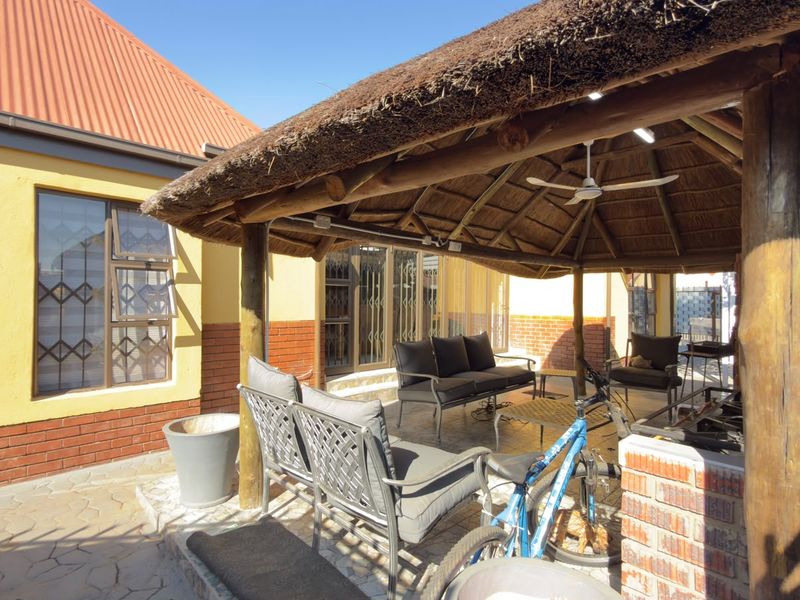 A covered outdoor patio area featuring a traditional thatch roof, timber support posts, and a built-in brick braai, ideal for outdoor entertaining.