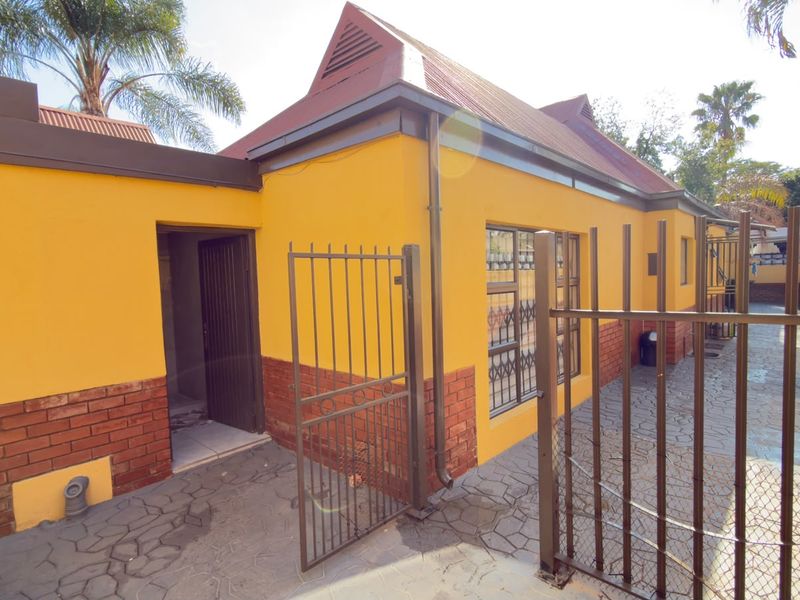 A rear exterior view of the property featuring a paved courtyard, brick and plaster facade, metal security gate, and security bars on the windows.