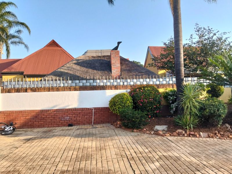Rear exterior view featuring a thatched roof structure, brick chimney, paved driveway, and established garden landscaping against a boundary wall.