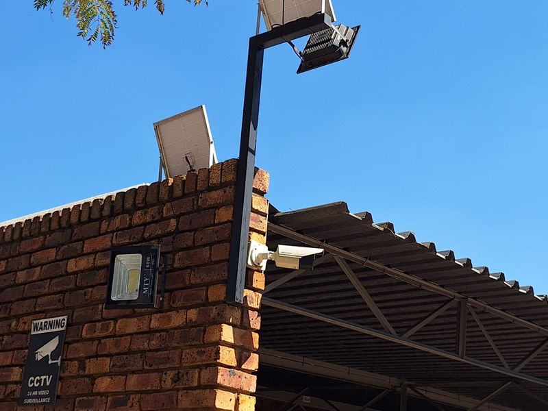 Exterior view of a brick wall featuring integrated solar-powered floodlighting and a CCTV security camera system under a corrugated metal roof structure.
