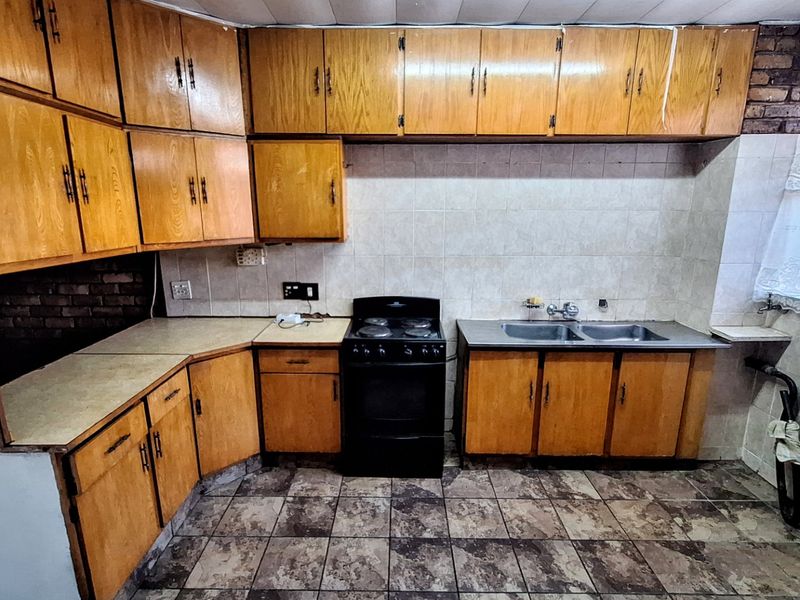 A dated kitchen featuring wooden cabinetry, laminate countertops, a double stainless steel sink, and a freestanding stove with tiled backsplash and flooring.