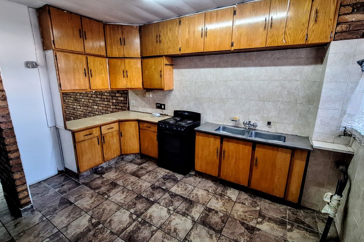 A dated kitchen featuring wooden cabinetry, tiled splashbacks, and a double basin sink, with an exposed brick feature wall and tiled flooring.