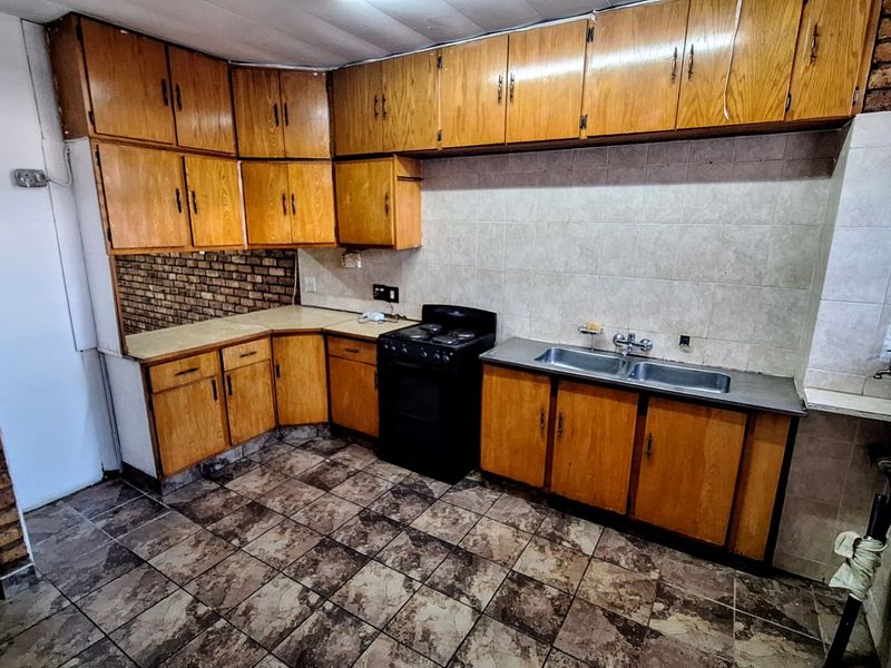 A dated kitchen featuring wooden cabinetry, tiled splashbacks, and a double basin sink, with an exposed brick feature wall and tiled flooring.