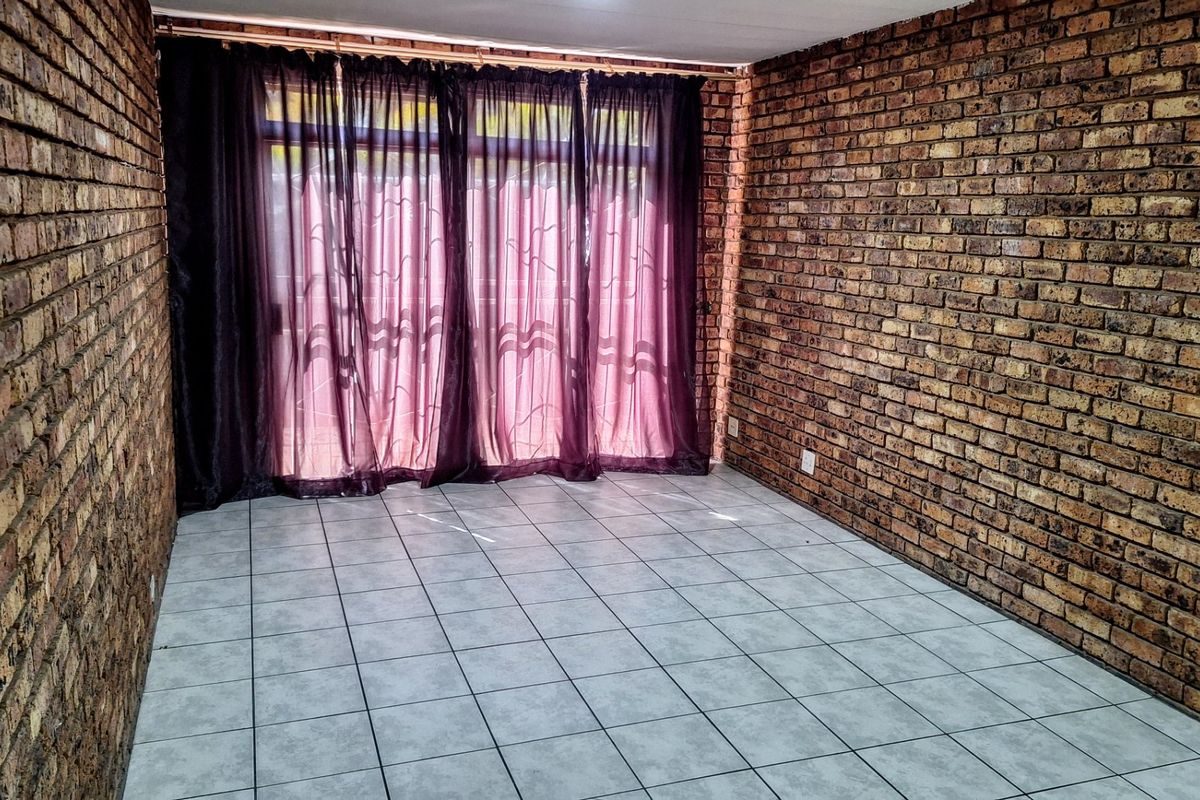 An empty living room featuring exposed brick walls and ceramic floor tiles, with a large sliding glass door providing natural light.